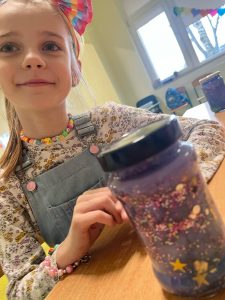 Uczennica klasy pierwszej siedzi przy stoliku. Przed nią stoi słoik wypełniony kolorowymi elementami imitującymi kosmos.