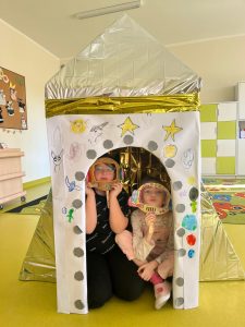 Dwoje uczniów klasy pierwszej pozuje do wspólnego zdjęcia siedząc wewnątrz makiety rakiety. Dzieci trzymają przed twarzą wycięte z papieru ramki w kształcie hełmów astronałtów.