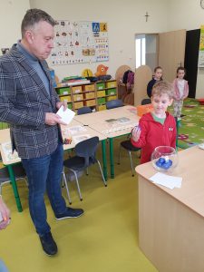 Uczeń oddziału przedszkolnego stoi przy przewodniczącym Rady Rodziców. Chłopiec trzyma w dłoni niebieską kulkę, która wylosował ze stojącej na biurku przed nim przezroczystej urny. Przewodniczący Rady Rodziców trzyma w dłoniach przed sobą koperty i przygląda się chłopcu.