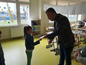 Przewodniczący Rady Rodziców przekazuje uczennicy klasy drugiej kopertę z nagrodą finansową. Dziewczynka uśmiecha się przejmując kopertę. Zdjęcie wykonane z profilu w sali lekcyjnej.