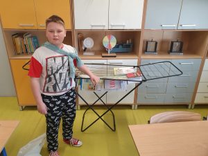 Uczeń klasy czwartej pozuje do zdjęcia stojąc przy suszarce do prania ustawionej na tle szafek w sali matematycznej. Na suszarce rozwieszone są podręczniki ucznia a chłopiec jedną ręką opiera się o nią.
