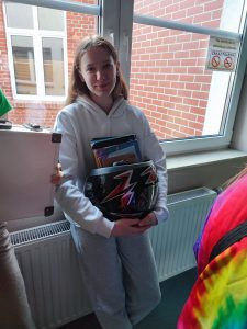 Uczennica klasy ósmej pozuje do zdjęcia trzymając w dłoniach przed sobą plastikowe wiaderko wypełnione książkami.