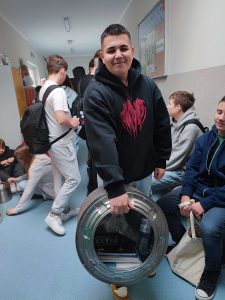 Uczeń lasy siódmej pozuje do zdjęcie trzymając w wyciągniętej ręce bęben od pralki, w którym umieszczone są podręczniki. w tle widoczni są inni uczniowie przebywający na szkolnym korytarzu.