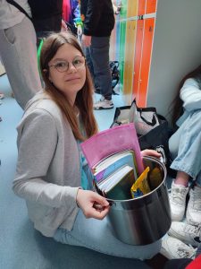 Uczennica klasy siódmej siedzi na podłodze. W dłoniach przed sobą trzyma garnek, w którym umieszczone są zeszyty i podręczniki.