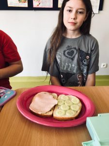 Uczennica klasy 4 siedzi przy stole. Przed dziewczynką na różowym talerzu leżą dwa kawałki chleba - na jednym z nich leży plasterek szynki a na drugim plasterki ogórka.