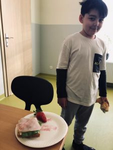 Uczeń klasy 4 stoi w niewielkiej odległości od stołu, w którego rogu ustawiono talerz z leżącą na nim kanapką przykrytą szynką oraz truskawką..