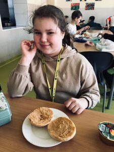 Uczennica klasy 4 siedzi przy stole. Dziewczynka uśmiecha się podpierając swoją dłoń o policzek. Przed nią na talerzu leży rozkrojona na pół bułka.