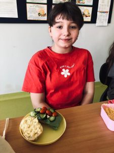 Uczennica klasy 4 siedzi przy stole. Przed dziewczynka na stole ustawiony jest plastikowy talerzyk z dwiema połówkami bułki. Na jednej widać twarożek, a na drugiej ładnie ułożone zielone i czerwone warzywa.