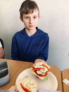 Uczeń klasy 4 siedzi przy stole. Na wyciągniętej nad nim dłoni trzyma kanapkę z dużą ilością warzyw. Chłopiec trzyma dłoń nad papierowym talerzu, na którym leży druga połowa bułki.