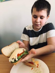 Uczeń klasy 4 siedzi przy stole. Chłopiec trzyma w dłoniach przed sobą kanapkę. W jednej dłoni trzyma jej spód z ułożonymi pod warstwą sera warzywami, a w drugiej kawałek chleba, którym lekko przytrzymuje spodnią część.