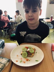 Uczeń klasy 4 siedzi przy stole. Przed chłopcem na stole leży na papierowym talerzu kanapka przybrana warzywami. W tle widać innych uczniów siedzących za stołem za nim.