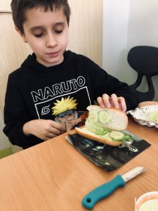 Uczeń klasy 4 siedzi przy stole. Uczeń jedną dłonią podtrzymuje otwartą kanapkę pokazując ułożone w jej wnętrzu zielone warzywa. Dolna część kanapki ułożona jest na serwetce spoczywającej na stole.