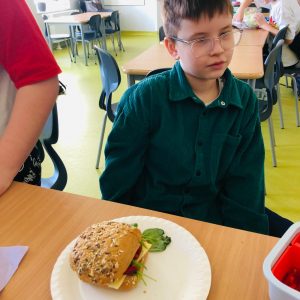 Uczeń klasy 4 siedzi przy stole. Przed chłopcem na papierowym talerzu leży złożona kanapka przygotowana z bułki z ziarnami.