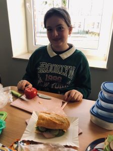 Uczennica klasy 4 siedzi przy stole. Dziewczynka uśmiecha się i ma oparte o stół dłonie. Przed nią na desce do krojenia leży pomidor oraz noże, a przed deską ustawiony jest papierowy talerzyk ze złożoną kanapką.