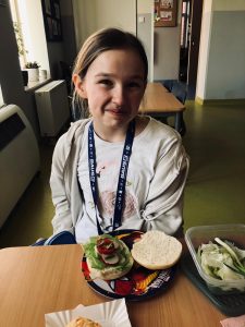 Uczennica klasy 4 siedzi przy stole. Dziewczynka uśmiecha się z zaciśniętymi ustami. Przed nią na talerzu leżą dwie połówki kanapki, z czego jedna przybrana jest warzywami.