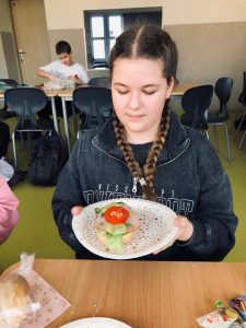 Uczennica klasy 4 siedzi przy stole. Dziewczynka w dłoniach przed sobą trzyma papierowy talerz w kolorowe kropki, na którym ułożona jest kanapka z pomidorem ozdobiona od góry plasterkami ogórka i pomidora. Dziewczynka patrzy na swoją kanapkę. W tle widoczny jest pracujący przy stole inny uczeń.