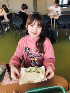 Uczennica klasy 4 siedzi przy stole. Dziewczynka w dłoniach przed sobą trzyma papierowy talerzyk, na którym ułożona jest połowa rogala ozdobiona krążkami z ogórka. Obok niej leży druga jej połowa.. W tle za dziewczynką widoczni są uczniowie pracujący nad swoimi kanapkami przy innych stołach.