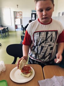 Uczeń klasy 4 stoi przy stole. Przed chłopcem na papierowym talerzu leży kanapka, której górną część chłopiec przytrzymuje jedną dłonią, by pokazać jej wnętrze. Na jej górze ułożone zostały duże kawałki pomidora. Chłopiec w drugiej dłoni trzyma drewniane sztućce.
