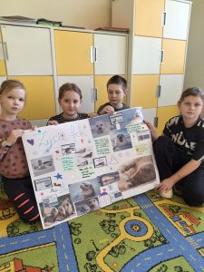 Czworo uczniów klasy drugiej pozuje do zdjęcia siedząc na dywanie i trzymając przed sobą przygotowany przez siebie plakat zatytułowany "Życie fok". Plakat stworzony jest z różnych zdjęć fok oraz odręcznie wypisanych na ich temat ciekawostek.