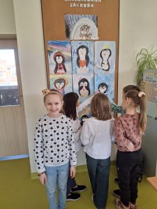 Uczennica klasy drugiej szeroko uśmiecha się do zdjęcia. Za nią stoi zwróconych do niej plecami czworo jej kolegów, którzy wskazują palcami, na wykonane odręcznie rysunki pingwinów umieszczone na tablicy korkowej.