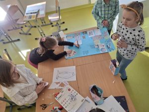 Czworo uczniów klasy drugiej pracuje wspólnie nad plakatem dotyczącym pingwinów. Jedna z dziewczynek zapisuje na nim informacje dotyczące życia tych zwierząt. Na stole pomiędzy uczniami poza plakatem leżą przybory do rysowania i wycinania.