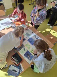 Przy stole na którym leżą przybory do rysowania i wycinanie czworo uczniów pracuje nad plakatem zatytułowanym "wieloryby". Jedna z dziewczynek pochyla swoje ciało nad plakatem zapisując na nim informacje dotyczące wielorybów.