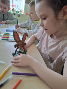 Uczennica oddziału przedszkolnego siedzi przy stole. Dziewczynka trzyma w dłoniach figurkę króliczka ze sklejki, którą ozdabia przy użyciu zielonego pisaka. Obok niej widać dwoje innych uczniów kolorujących pisanki wydrukowane na papierze.