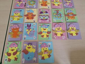 Zdjęcie ułożonych obok siebie kartek wielkanocnych wykonanych przez uczniów oddziału przedszkolnego. Na kolorowym papierze uczniowie wykleili z kolorowych pianek zajączki, kurczaczki i pisanki.
