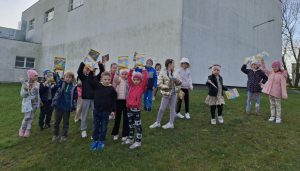 Uczniowie oddziału przedszkolnego stoją na trawniku przed budynkiem szkoły. Dzieci podnoszą w górę książeczki z nadrukiem wielkanocnym na okładce.