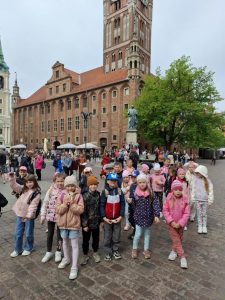Uczniowie biorący udział w wyjeździe pozują do wspólnego zdjęcia stojąc na rynku w Toruniu. Za nimi widoczny jest pomnik Mikołaja Kopernika oraz ratusz.