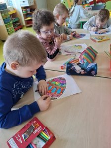 Czworo uczniów z oddziału przedszkolnego siedzi przy wspólnym stole. Dzieci kolorują pisakami pisanki nadrukowane na białych kartkach papieru. Przed dziećmi leżą piórniki i pudełka z pisakami.