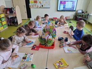 Uczniowie z oddziału przedszkolnego siedzą przy wspólnym stole. Dzieci kolorują pisakami pisanki nadrukowane na białych kartkach papieru. Przed nimi leżą piórniki i pudełka z pisakami i kredkami. Na środku stołu ustawione są dwie duże kuwety a w nich wielkanocne ozdoby. Z doniczek w kształcie połowy skorupki jajka wyrasta trawka, w która włożone są patyczki zakończone okręgami wyciętymi z papieru z rysunkami zajączków i kurczaczków.