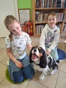 Dwie uczennice klasy drugiej kucają na kolanach i głaszczą pieska siedzącego przed nimi. Jedna z z dziewczynek patrzy pod kątem w stronę pieska a druga prosto w stronę obiektywu.