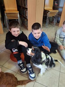 Dwóch uczniów klasy drugiej siedzi na podłodze biblioteki w Ostaszewie. Chłopcy patrzą w stronę pieska siedzącego przed nimi. Jeden z nich wyciąga swoją dłoń nad grzbiet zwierzaka.