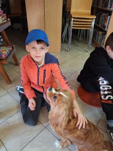 Uczeń klasy drugiej siedzi na podłodze. Chłopiec wyciąga jedną z rąk głaskając pieska siedzącego przed nim. Chłopiec patrzy w stronę obiektywu a piesek ma pysk zwrócony w jego kierunku.