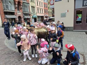 Uczniowie biorący udział w wycieczce pozują do wspólnego zdjęcia stojąc bądź kucając wokół ustawionego na rynku w Toruniu pręgierza w kształcie osiołka wykonanego z metalu.
