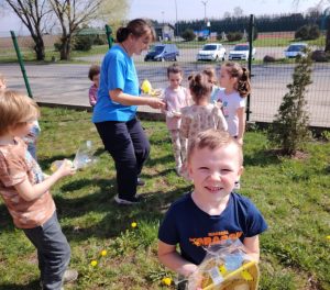 Uczniowie oddziału przedszkolnego wraz z wychowawczynią stoją na trawniku przed szkolnym ogrodzeniem. Kobieta rozdaje dzieciom upominki umieszczone w przezroczystej folii. Na pierwszym planie widoczny jest chłopiec trzymający w dłoni otrzymany upominek i patrzący w stronę obiektywu.