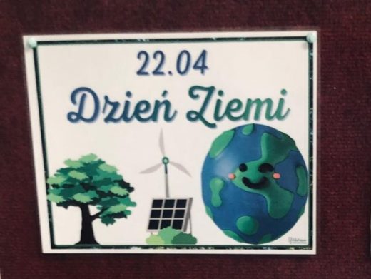 Na czerwonym tle kartka papieru z nadrukowaną czarną ramką. Wewnątrz ramki napisy: "22.04 Dzień Ziemi". Poniżej umieszczone grafiki drzewa, wiatraka słonecznego i paneli oraz ulepionej z plasteliny Ziemi z przyklejonymi oczkami i uśmiechem.