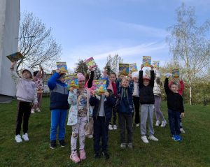 Uczniowie oddziału przedszkolnego stoją na trawniku przed budynkiem szkoły. Dzieci podnoszą w górę książeczki z nadrukiem wielkanocnym na okładce.