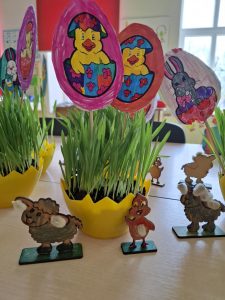 Zdjęcie wykonanych przez uczniów dekoracji wielkanocnych. Na stole ustawione są żółte doniczki przypominające połówki skorupek jajka, z których wyrasta trawka. W doniczkach umieszczone zostały patyczki zakończone wyciętymi z papieru jajkami. Na jajkach tych są nadrukowane i pokolorowane przez dzieci kurczaczki i zajączki. Przed doniczkami ustawione zostały figurki ze sklejki również pokolorowane przez uczniów. Figurki przedstawiają baranki i kurczaczka i dodatkowo ozdobione są baziami.