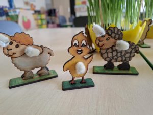 Zdjęcie trzech figurek wykonanych ze sklejki i ozdobionych baziami. Figurki przedstawiają dwa baranki i kurczaczka. Ich podstawki są pomalowane na zielono.