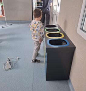 Uczeń oddziału przedszkolnego w bluzie z postaciami z bajki stoi przy zestawie pojemników do segregacji odpadów oznaczonych kolorami: zielonym, żółtym, niebieskim i czarnym. Obok na podłodze leży żelazko, a w tle widać automat z przekąskami i osobę dorosłą przy drzwiach.