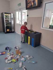 Uczeń oddziału przedszkolnego w czerwonej bluzie z postacią Spider-Mana wrzuca odpad do pojemnika do segregacji śmieci na szkolnym korytarzu. Za nim na podłodze leży rozrzucone różnorodne śmieci, a w tle widać automat z przekąskami, drzwi i telewizor.