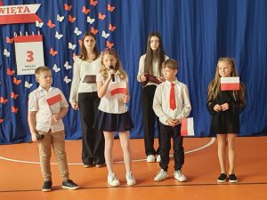Czoro uczniów edukacji wczesnoszkolnej stoi w rzędzie trzymając w dłoniach polskie flagi. Za nimi widać uczennice prowadzące apel. W tle na niebieskiej tkaninie zawieszone są dekoracje w kolorach białym i czerwonym. Na zdjęciu widoczne są wycięte z papieru motyle oraz dekoracja przypominająca kartkę z kalendarza z napisem "3 ŚWIĘTO KONSTYTUCJI".