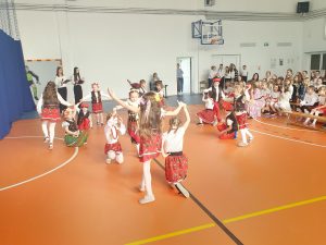 Uczniowie oddziału przedszkolnego ubrani w stroje krakowiaków tańczą na sali gimnastycznej. Część z dzieci kuca podczas, gdy ich koleżanki okrążają ich z wyciągniętymi w górę ramionami. W tle widać siedzącą na sali widownię.