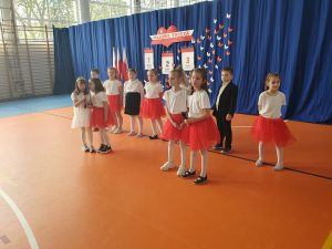 Uczniowie klasy pierwszej stoją na sali gimnastycznej podczas występu na apelu. Uczennice ubrane są w białe koszulki i czerwone lub czarne spódniczki, a chłopcy w garnitury. Część z dzieci ustawiona jest w jednym rzędzie a przed nimi stoją w dwóch dwuosobowych grupach ich koleżanki. W tle widoczny jest niebieski materiał na którym zawieszona została dekoracja wykonana w kolorach białym i czerwonym - - napis "MAJOWE ŚWIETA" oraz trzy dekoracje przedstawiające kartki z kalendarza ponumerowane od 1 do 3 z wypisanymi pod nimi nazwami świąt. całość uzupełniają białe i czerwone motyle wycięte z papieru.