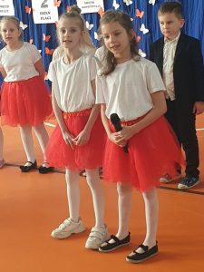 Dwie uczennice u=edukacja wczesnoszkolnej ubrane w identyczne biało - czerwone stroje stoją obok siebie na sali gimnastycznej. Jedna z nich trzyma w dłoniach przed sobą mikrofon. W tle widać innych uczniów stojących w rzędzie. oraz fragment dekoracji sali.