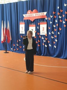 Uczennica klasy trzeciej stoi sama na scenie trzymając w dłoniach przed sobą mikrofon. Dziewczynka stoi na tle rozwieszonej niebieskiej tkaninie na której umieszczono dekoracje wykonaną w kolorach białym i czerwonym - napis "MAJOWE ŚWIETA" oraz trzy dekoracje przedstawiające kartki z kalendarza ponumerowane od 1 do 3 z wypisanymi pod nimi nazwami świąt. całość uzupełniają białe i czerwone motyle wycięte z papieru.
