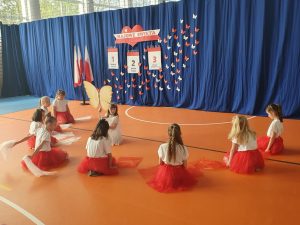 Uczennice ze szkolnego kółka tańca siedzą w czerwonych spódniczkach tworząc półokrąg na podłodze sali gimnastycznej. Po środku półokręgu kuca ich koleżanka w białej sukience trzymająca kij na którym umieszczone są wycięte z tektury skrzydła motyla. W tle widoczna jest niebieska tkanina, na której umieszczona dekoracje w białym i czerwonym kolorze - - napis "MAJOWE ŚWIETA" oraz trzy dekoracje przedstawiające kartki z kalendarza ponumerowane od 1 do 3 z wypisanymi pod nimi nazwami świąt. całość uzupełniają białe i czerwone motyle wycięte z papieru.