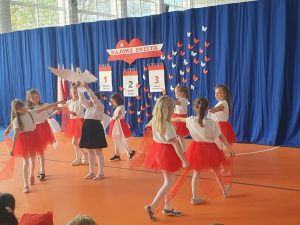 Uczennice ze szkolnego kółka tańca ubrane w białe bluzki i czerwone spódniczki wirują wokół stojącej po środku koleżanki trzymającej nad głową wyciętej z papieru postaci orła. W tle widoczna jest umieszczona na niebieskim materiale dekoracja wykonana w czerwonym i białym kolorze - - napis "MAJOWE ŚWIETA" oraz trzy dekoracje przedstawiające kartki z kalendarza ponumerowane od 1 do 3 z wypisanymi pod nimi nazwami świąt. całość uzupełniają białe i czerwone motyle wycięte z papieru.